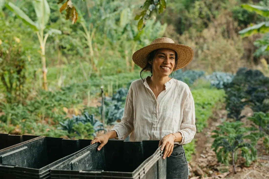 Produtora sorri ao lado de caixas de colheita em uma horta diversificada.